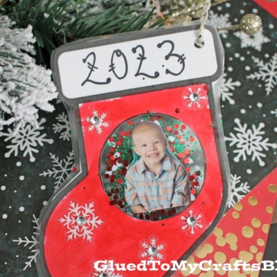 A handmade stocking photo ornament with 2023 on top, featuring your child’s picture in the center. The red stocking is decorated with snowflakes and displayed on festive wrapping paper.