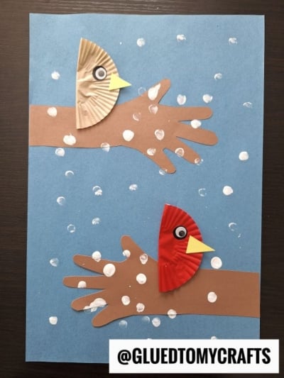 A craft showing two handprints made from brown paper as tree branches on blue paper. Each branch has a cupcake liner bird with a googly eye and yellow beak. Q-tip painted winter tree snow is shown with white dots. @GLUEDTOMYCRAFTS is at the bottom.