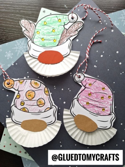 Three handmade Cupcake Liner Christmas Gnome ornaments with colorful hats and cut-out noses, crafted from paper and cupcake liners, rest on festive patterned paper. Each gnome ornament has a string for easy hanging.