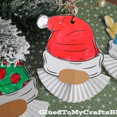 Three handmade Cupcake Liner Christmas Gnome ornaments made from colored paper and white cupcake liners are arranged on green patterned paper, with festive decorations nearby.