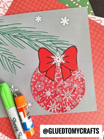 A handmade Christmas card features a red Cupcake Liner Ornament adorned with white snowflakes and a red paper bow, set against a gray background with drawn green pine needles. Supplies are nearby.