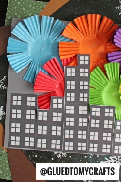A cityscape collage features black paper buildings with white windows and vibrant cupcake liner fireworks bursting in the night sky. @GLUEDTOMYCRAFTS is written at the bottom.