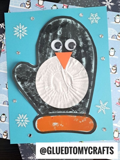 A paper penguin mitten craft made with a black mitten shape, white cupcake liner belly, orange paper beak and feet, googly eyes, and snowflake stickers on a blue background. Text: @GLUEDTOMYCRAFTS.