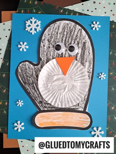 A handmade paper penguin mitten craft on blue paper features a black mitten shape, googly eyes, an orange beak, a white cupcake liner belly, and snowflake stickers, with @GLUEDTOMYCRAFTS printed at the bottom.