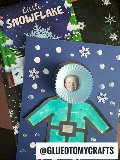 A children’s snow-themed craft features a child’s photo in a blue cupcake liner as the head of a paper winter coat snowsuit, next to a “Little Snowflake” book. Text at the bottom reads @GLUEDTOMYCRAFTS.