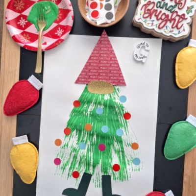 A Christmas craft project features a gnome made from paper and green fork painted details, decorated with colorful dot stickers, surrounded by felt Christmas lights, festive plates, cutlery, and a “Merry and Bright” sign.