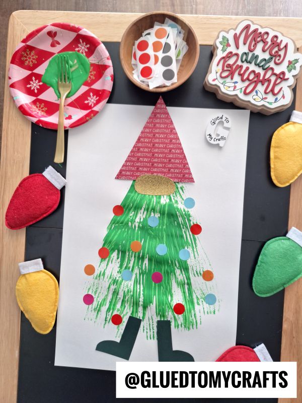 A Christmas craft project features a gnome made from paper and green fork painted details, decorated with colorful dot stickers, surrounded by felt Christmas lights, festive plates, cutlery, and a “Merry and Bright” sign.