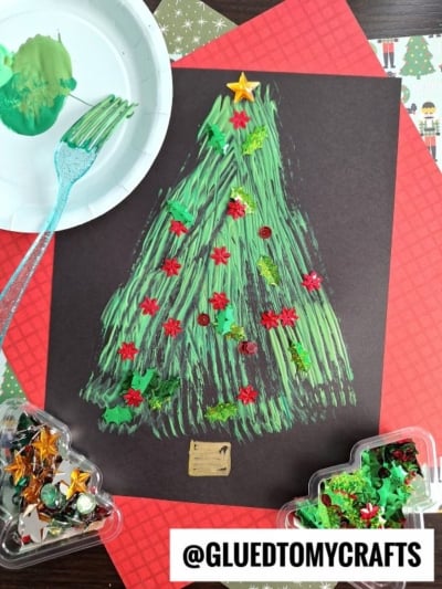 A fork painted Christmas tree is textured with green paint and fork strokes on black paper, decorated with red and gold star-shaped sequins and green glitter. Craft supplies and a green-painted fork are nearby.