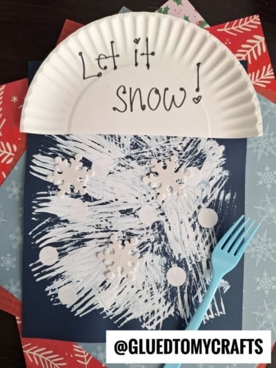 A paper plate with Let it snow! sits above a winter craft made with fork painted white snowflakes and circle cutouts on dark paper. A blue plastic fork and snowflake-patterned papers are nearby.
