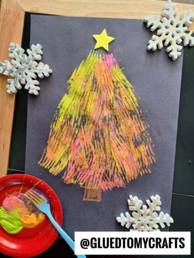 A child’s fork painted Christmas tree in yellow, pink, and orange on black paper, topped with a yellow star, is surrounded by painted wooden snowflakes and a red plate with paint and a blue brush. Text reads @GLUEDTOMYCRAFTS.