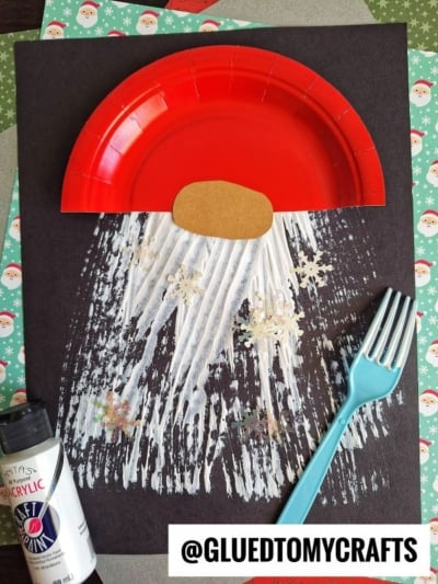 A Santa craft made with a red paper plate as a hat, a brown oval nose, and a fork painted white beard dusted with snowflake glitter on black paper; a blue fork and white paint bottle are beside it.