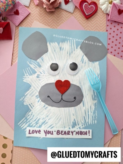 A bear face made from gray and white paper, googly eyes, a red glitter heart nose, and a fork painted blue, with a note saying Love You Beary Much! on a colorful, crafty background.