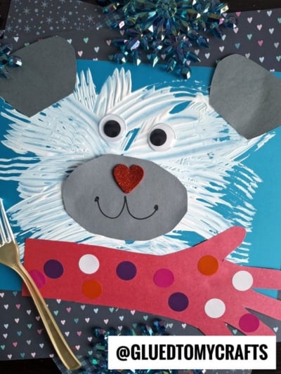 A winter polar bear craft made with fork painted white fur, large googly eyes, gray paper ears and snout, a red glitter heart nose, and a red paper mitten with polka dots, all arranged on a blue background.