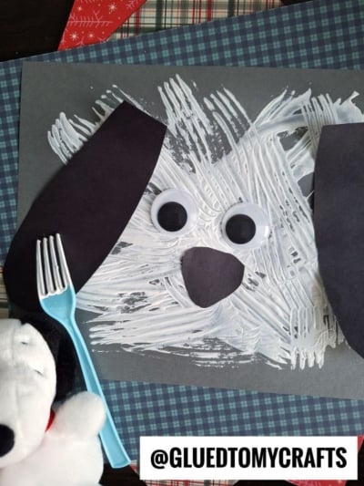 A textured dog face is fork painted with white paint on dark paper, featuring two large googly eyes, black paper ears, and a nose. A blue fork and plush dog toy are beside the artwork. @GLUEDTOMYCRAFTS is displayed.