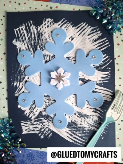 A large blue foam snowflake and a small white snowflake are glued to dark paper with fork painted white strokes, a plastic fork, and festive decorations nearby. The text @GLUEDTOMYCRAFTS appears at the bottom.