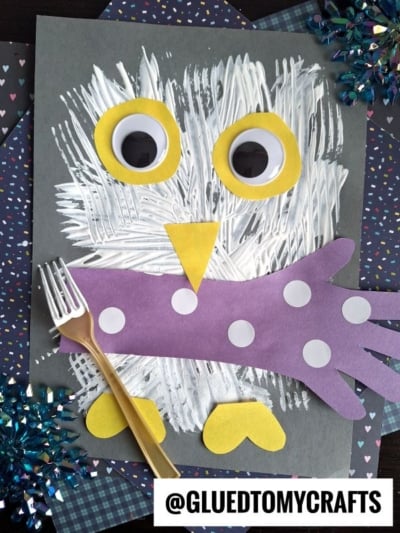 A handmade owl craft on black paper, featuring a body fork painted with white streaks, two large yellow and white eyes, a yellow triangle beak, purple wings with white dots, and yellow feet, next to a plastic fork.