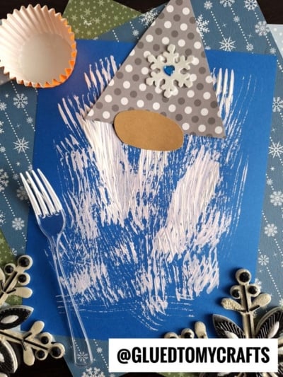 A winter gnome craft on blue paper, featuring a polka-dot hat, brown nose, and a fork painted white beard. Decorative snowflake papers, a cupcake liner, and metallic embellishments surround the artwork.