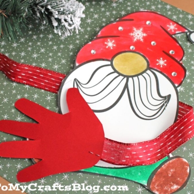 A handmade gnome craft features a red hat, white beard, and wide red arms in a gnome handprint hug. The background includes green patterned paper and holiday decor.