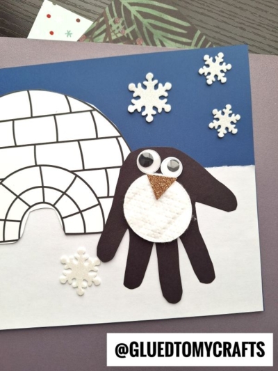 A handprint penguin craft features a black penguin made from a handprint cutout, with googly eyes and a gold beak, beside an igloo and paper snowflakes on a blue and white background.
