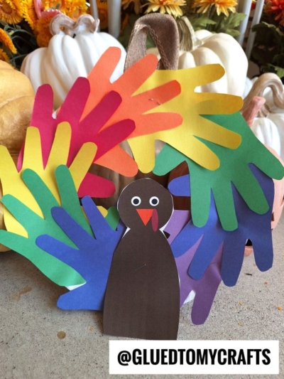 A colorful handprint feather turkey craft made from layered paper in red, yellow, orange, green, blue, and purple, with a brown paper body and face, displayed among white pumpkins and flowers. @GLUEDTOMYCRAFTS is written below.