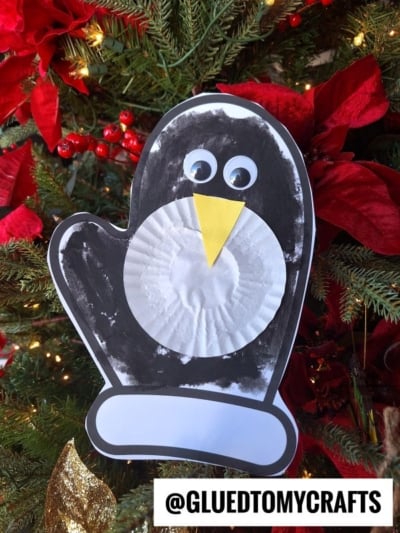 A handmade paper penguin mitten ornament, crafted with a black mitten-shaped paper, a white cupcake liner belly, googly eyes, and a yellow beak, hangs on a Christmas tree adorned with red flowers and greenery.