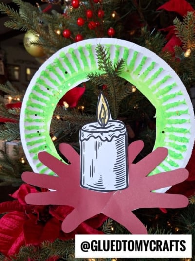 A handmade advent wreath craft made from a green-painted paper plate, red paper cutout hands, and a drawing of a candle, hanging on a decorated Christmas tree. Text reads @GLUEDTOMYCRAFTS.