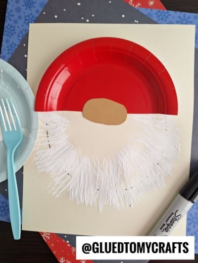 A Santa craft made with a red paper plate for the hat, a tan oval for the nose, and a fork painted white beard on cream paper. A black marker and plastic utensils are nearby. @GLUEDTOMYCRAFTS is written at the bottom.