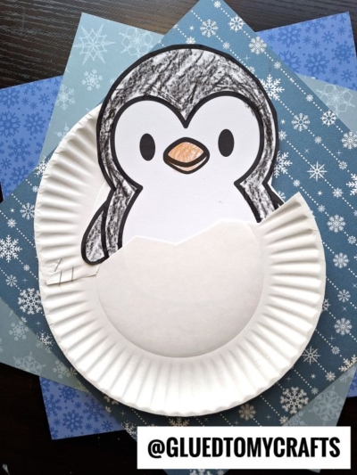 A paper plate hatching penguin craft featuring a cute black and white penguin emerging from an eggshell, set against blue paper with snowflake patterns. @GLUEDTOMYCRAFTS is at the bottom.