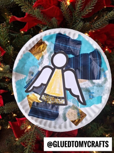 A tissue paper angel craft made with blue, gold, and white tissue pieces and a paper angel cutout, displayed on a Christmas tree with red ribbon and lights. @GLUEDTOMYCRAFTS is written at the bottom.