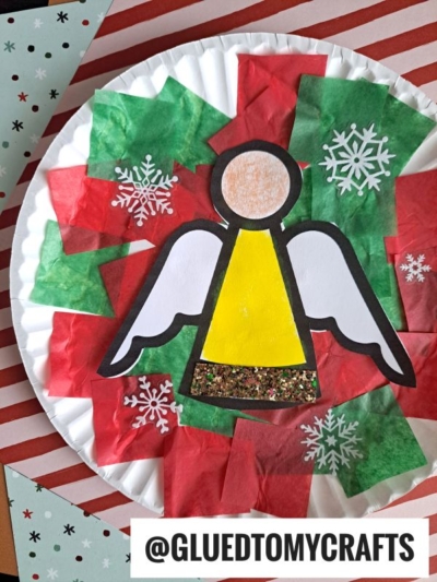 A paper plate Christmas craft featuring a tissue paper angel with a yellow dress and glitter, white wings, and a round face, surrounded by red and green tissue paper squares and white snowflake stickers.