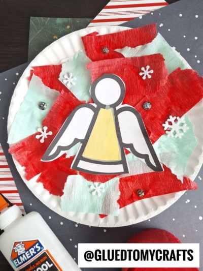 A paper plate decorated with red and mint green tissue paper, white snowflakes, a tissue paper angel cutout, and glitter accents. Elmer’s glue and craft materials are nearby. @GLUEDTOMYCRAFTS is written at the bottom.