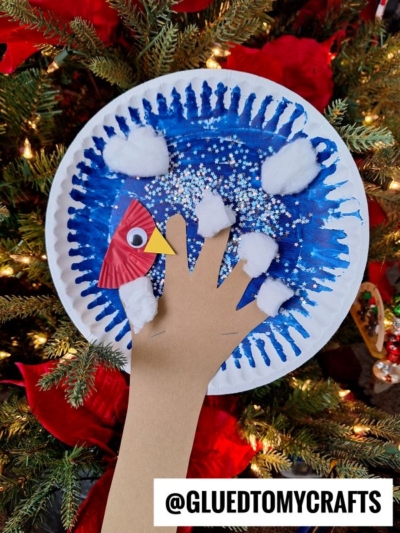 A paper plate winter tree craft features a blue, snow-dotted plate with cotton ball snow, a brown paper hand as the tree branch, and a red bird from cupcake liners. Christmas tree branches add festive background charm.