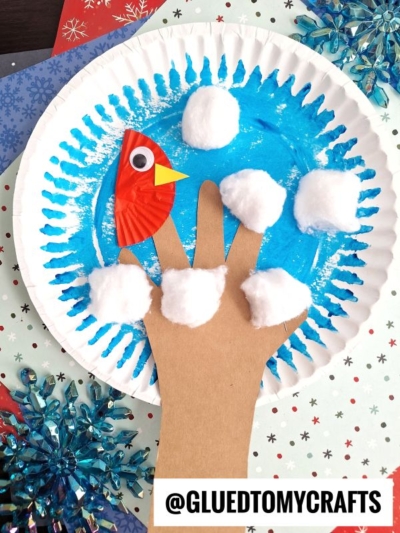A paper plate winter tree craft: a blue-painted plate with a brown cutout hand as the tree, cotton ball snow, and a red cupcake liner bird with a googly eye and yellow beak. Winter-themed paper snowflakes decorate the background.