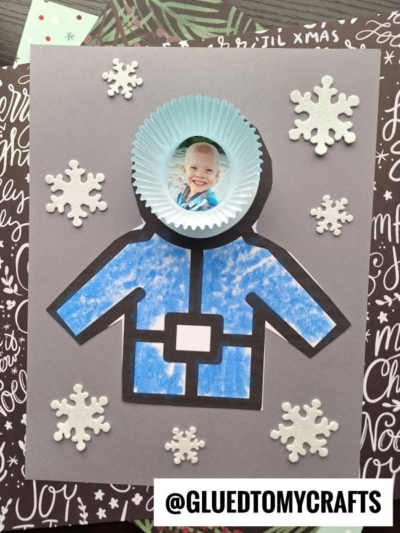 A handmade astronaut craft featuring a child’s photo in a blue paper cupcake liner as the helmet, with a drawn blue suit and paper winter coat, surrounded by white snowflake cutouts on a black and white background.