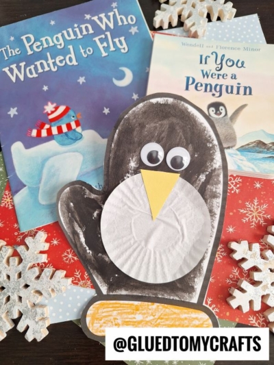 A handmade paper penguin mitten craft with googly eyes and a yellow beak is placed on red snowflake paper, surrounded by two children's books about penguins, paper snowflakes, and a @gluedtomycrafts label.