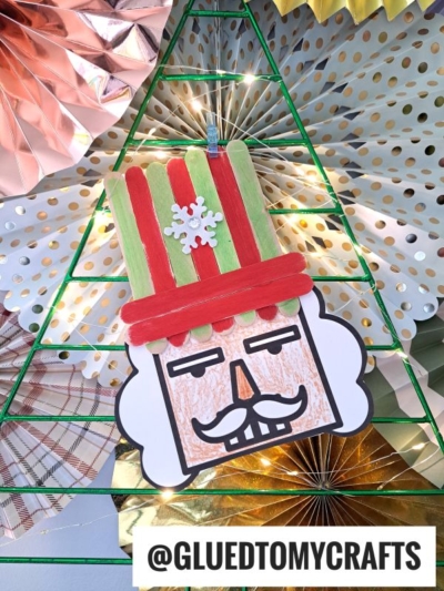 A handmade nutcracker craft with a red and green striped nutcracker hat, a snowflake decoration, and a mustache is displayed on a wire-frame Christmas tree with metallic paper fans in the background. @GLUEDTOMYCRAFTS is captioned at the bottom.