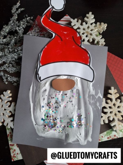 A handmade puffy paint Santa craft with a red hat, an oval nose, and a sparkly white beard on gray paper. Snowflake decorations and craft supplies surround the project. @GLUEDTOMYCRAFTS is at the bottom.