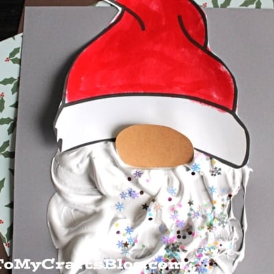 A puffy paint Santa Claus craft featuring a paper hat, a brown oval face, and a fluffy white beard made from shaving cream and glitter, displayed on gray paper with holiday-themed sheets underneath.