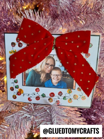 A handmade photo ornament with a red bow hangs on a pink Christmas tree. Festive Q-Tip Painted Christmas Lights are drawn around the smiling woman and child in the photo, with the tag @gluedtomycrafts.