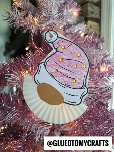 A Cupcake Liner Christmas Gnome with a pink hat and gold dots hangs on a pink Christmas tree. Handmade from paper, the ornament features a beard crafted from a cupcake liner and has @GLUEDTOMYCRAFTS labeled below.