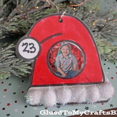 A red santa claus hat ornament, shaped like Santa’s hat with a round cutout for a child’s photo and labeled 23, hangs on a Christmas tree.