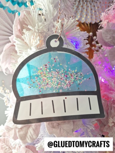A paper ornament shaped like a snow globe, filled with colorful glitter, hangs on a white and pink decorated Christmas tree—perfect beside a winter hat suncatcher. Text at the bottom reads @GLUEDTOMYCRAFTS.