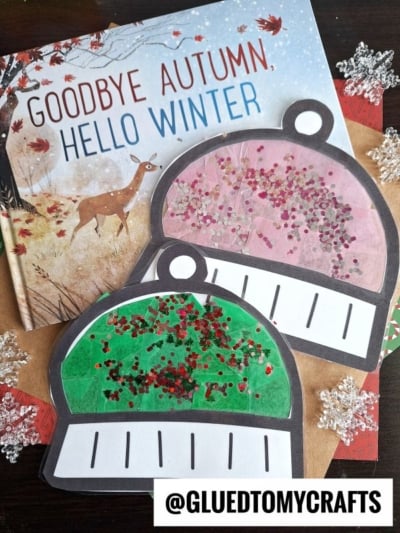 A book titled Goodbye Autumn, Hello Winter is shown with two paper snow globe crafts—one green, one pink—decorated with red glitter on top. Silver snowflake ornaments, a winter hat suncatcher, and a handle with @GLUEDTOMYCRAFTS are also visible.