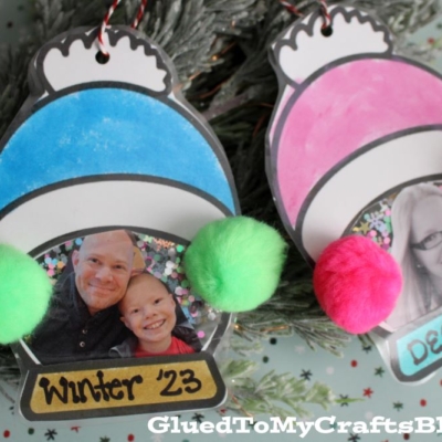 Handmade winter hat ornaments feature family photos and colorful pom-poms, labeled Winter 23. One green ornament shows a man and child; the pink winter hat ornament displays a woman. Pine branches and gift wrap add festive charm to the background.