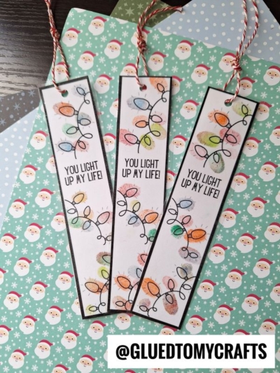 Three handmade Light Up My Life Bookmarks with colorful Christmas lights and the phrase You light up my life! are displayed on festive Santa-themed paper. Each bookmark has a red and white string tied at the top. @gluedtomycrafts text is at the bottom.