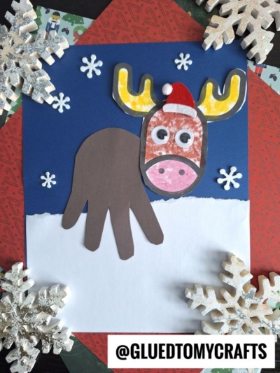 A handmade reindeer or handprint moose craft with a brown handprint body, decorated face, googly eyes, and antlers on blue paper with snowflakes. Large faux snowflakes border the image. @GLUEDTOMYCRAFTS is written at the bottom.