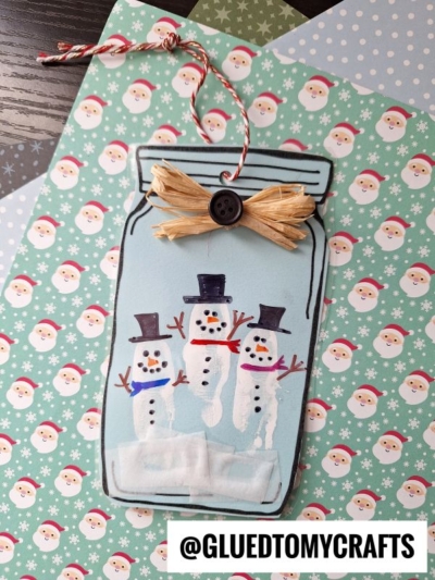 A handmade mason jar snowman card decorated with three snowmen made from white fingerprints, each with hats and scarves, a raffia bow with a black button, and festive Santa-themed paper in the background.