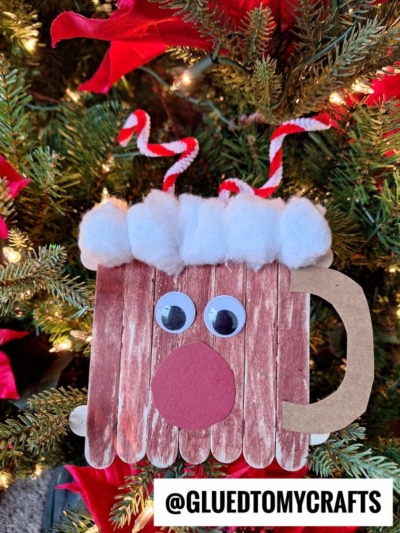A reindeer face ornament, inspired by a reindeer hot cocoa mug, is crafted from sticks, cotton balls, googly eyes, and a red nose. Red and white striped pipe cleaner antlers complete the look. @GLUEDTOMYCRAFTS is labeled at the bottom.