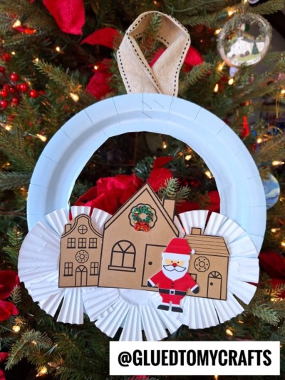 A handmade winter village wreath ornament crafted from a white paper plate, paper rosettes, and cutouts of houses and a Santa figure, hanging on a decorated Christmas tree. @GLUEDTOMYCRAFTS is written at the bottom.