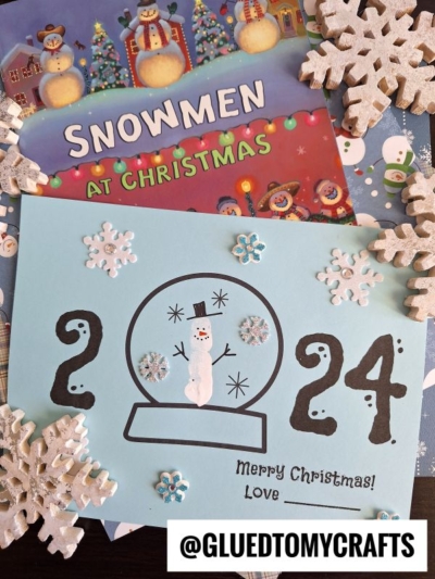 A blue 2023 snowman card with “Merry Christmas!” sits near a snow globe and festive snowflakes. Behind it is the book Snowmen at Christmas and the Instagram handle @GLUEDTOMYCRAFTS.